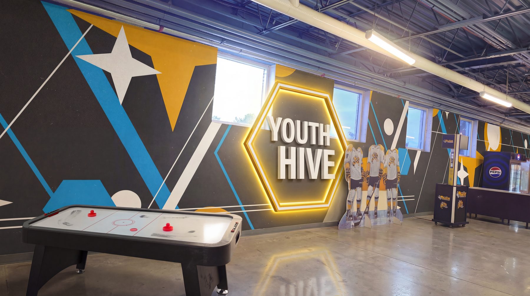 Youth Hive: a lit hexagon sign on a wall of bold yellow, blue and white shapes, with an air hockey table in front